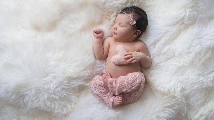 Sleeping, week old newborn baby girl wearing light pink, knitted pants and holding a tiny, heart shaped pillow. Shot in the studio on a white sheepskin rug.