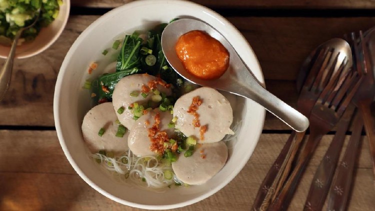 1177933432 Bakso Gepeng, a popular street food dish of flat meatballs soup. Served with rice noodles, green leafy vegetables, scallion and fried garlic. Accompanied with red chili sauce. The dish is placed on a rustic wooden table with a bowl of extra scallion.