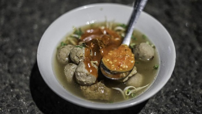 Bakso Gepeng, a popular street food dish of flat meatballs soup. Served with rice noodles, green leafy vegetables, scallion and fried garlic. Accompanied with red chili sauce. The dish is placed on a rustic wooden table with a bowl of extra scallion.