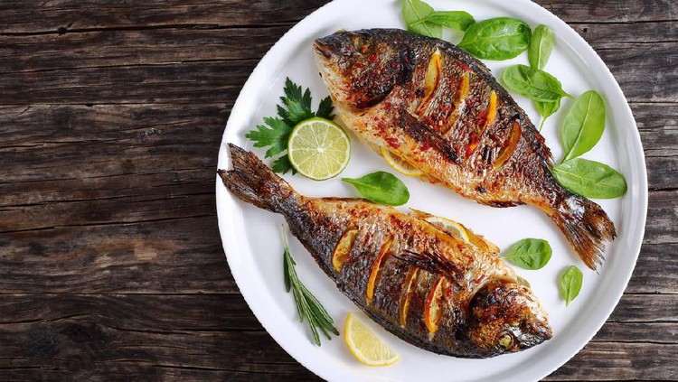 delicious roasted dorado or sea bream fish with lemon and orange slices, spices, fresh parsley and spinach on white platter on old dark wooden table view from above