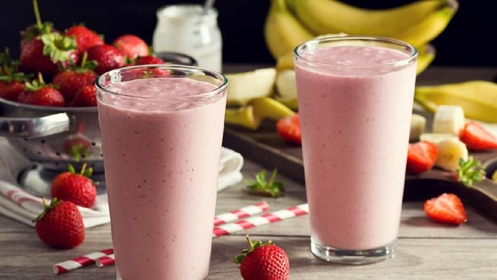 Two cold strawberry banana yogurt smoothies in pint glasses with striped straws. The berries and bananas are in the background in a colander and on a cutting board. A jar of yogurt is also in the background. Everything is on a faded wood picnic table.