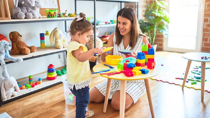Young beautiful teacher and toddler playing with dishes and cutlery toy at kindergarten