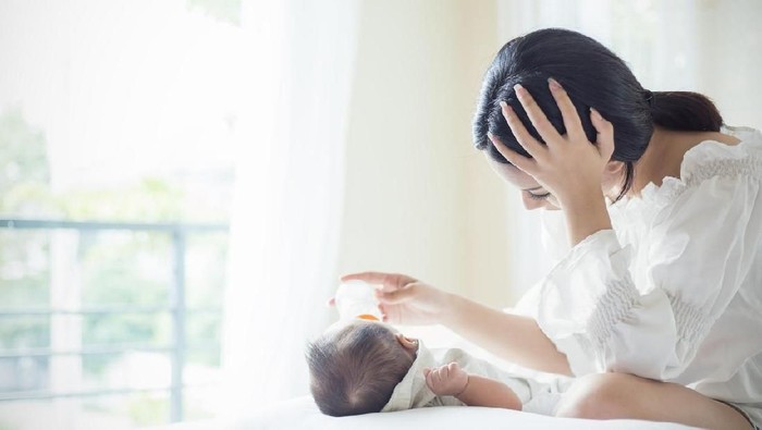 Asian Mother nursery feeding bottle of formula milk to newborn baby in bed suffering from post natal depression. Health care single mom motherhood stressful concept.
