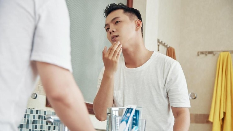 From below back view of man touching face skin looking at reflection of bathroom mirror at home