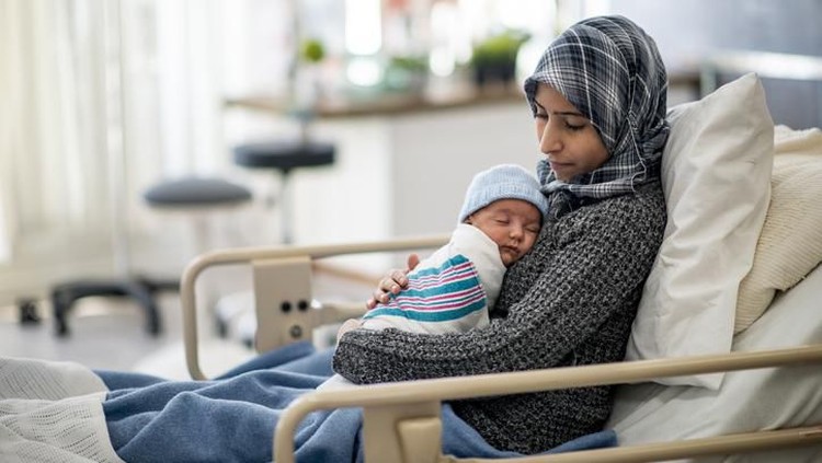 A Muslim woman is laying in a hospital bed. She is holding her newborn son on her lap. She is watching over her baby while he sleeps.