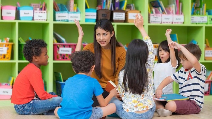 A multi-ethnic group of preschool students is sitting with their legs crossed on the floor in their classroom. The mixed-race female teacher is sitting on the floor facing the children. The happy kids are smiling and following the teacher's instructions. They have their arms raised in the air.