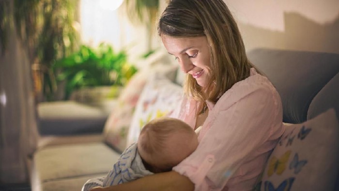 Young beautiful mother, breastfeeding her newborn baby boy at night, dim light. Mom breastfeeding infant
