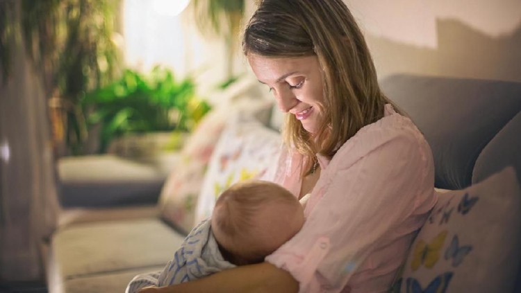 Young beautiful mother, breastfeeding her newborn baby boy at night, dim light. Mom breastfeeding infant