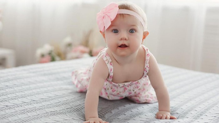 Charming beautiful lovely small female baby with plump cheeks creeps on bed, looks innocently on something, being naughty. Cute baby infant plays alone. Newborn kid crawls. Enthusiastic child
