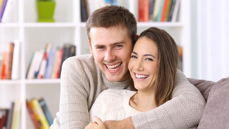 Front view portrait of a happy couple with perfect smile looking at you sititng on a couch in the living room at home in winter