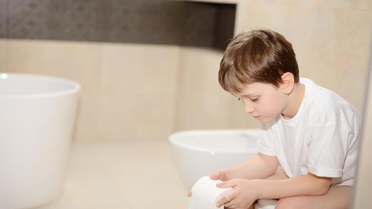 Little 7 years old boy sitting on toilet. Holding white toilet paper