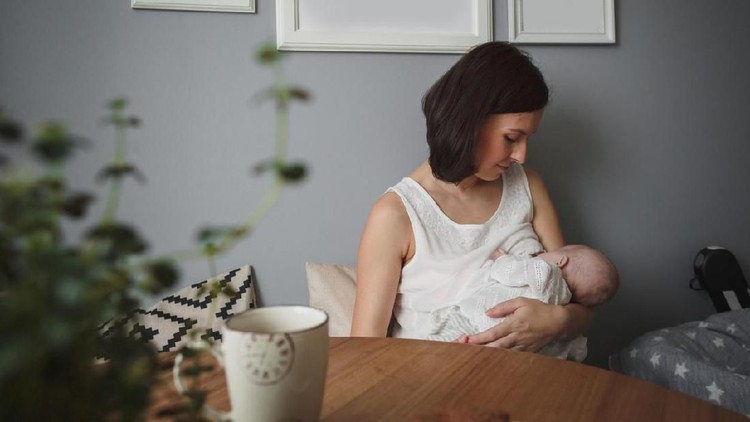Young beautiful woman is breastfeeding a little baby in a cozy room. On the table there is a white Cup and a green plant. On the wall hang a white empty picture frames. No images of nudity