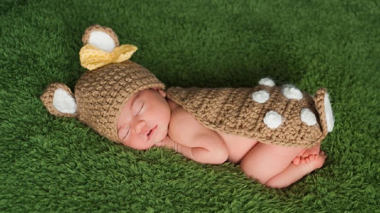 A newborn baby girl wearing a Whitetail deer / fawn costume. She is sleeping on a grass green fuzzy blanket.
