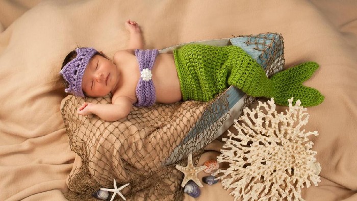 Newborn baby girl wearing a crocheted green and lavender colored mermaid costume. She is sleeping in a little boat on a studio set with sand colored fabric, fish net and sea shells.