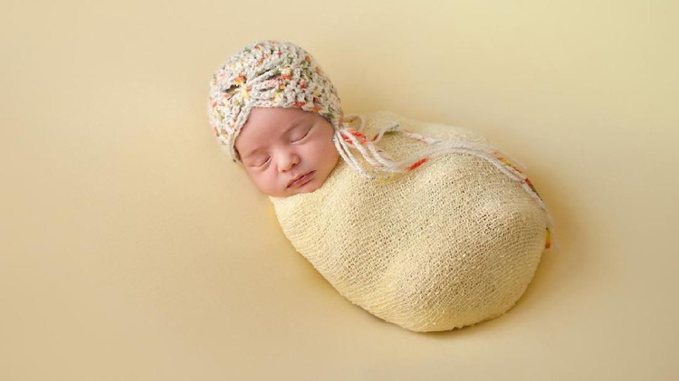 A portrait of a beautiful, two week old, newborn baby girl wearing a crocheted bonnet. She is swaddled and sleeping on yellow colored fabric.