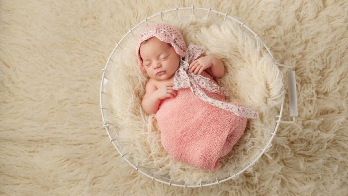 A portrait of a five week old newborn baby girl wearing a pink bonnet and sleeping in a wire basket. Shot from overhead.