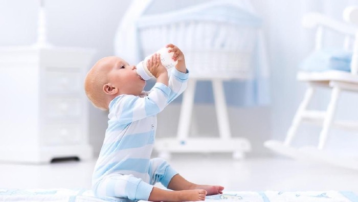 Adorable baby boy playing on a blue floor mat and drinking milk from a bottle in a white sunny nursery with rocking chair and bassinet. Bedroom interior with infant crib. Formula drink for infant.