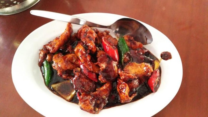Ayam Rica-Rica, the popular and traditional dish from Manado, chicken braised in spicy red chili paste. Two drumsticks are arranged on a small ceramic plate that has been lined with a banana leaf. Two lime halves and a lime leaf are used to garnish the dish. The plate is placed on a square bamboo plate with a pair of wooden cutlery tucked in. The bamboo plate is placed on a rustic wooden table.
