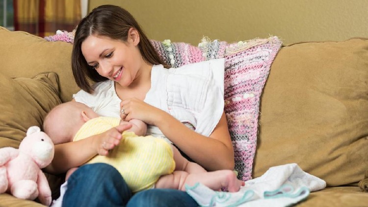 Young adult Caucasian woman is sitting on sofa at home in living room. She is breastfeeding her six month old baby daughter. Mother is smiling lovingly at baby while nursing her.