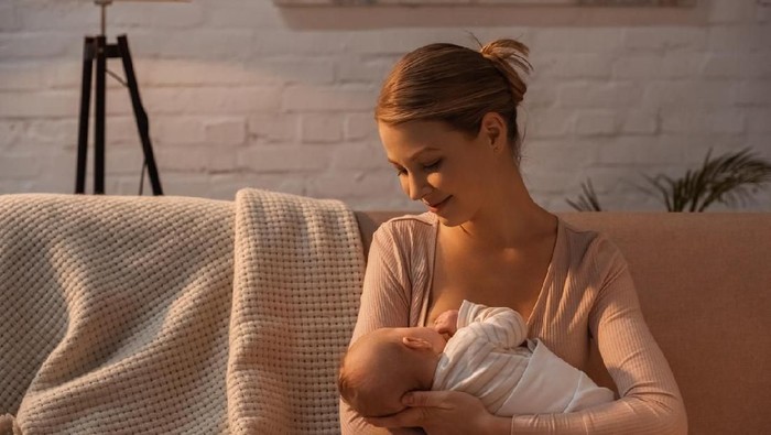 Young adult Caucasian woman is sitting on sofa at home in living room. She is breastfeeding her six month old baby daughter. Mother is smiling lovingly at baby while nursing her.