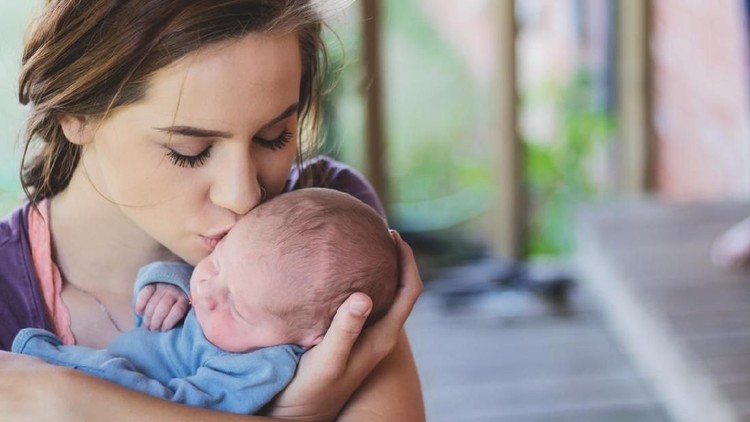 Young teenage Caucasian woman is mother to newborn baby boy. She kisses him on the cheek as she holds him lovingly in her arms.