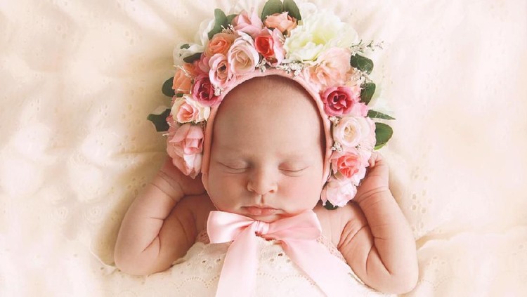 A newborn baby girl, fast asleep, wears a bonnet of flowers.