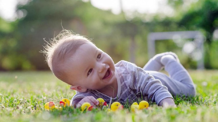 953026562 Cute funny laughing Caucasian baby boy learning to crawl, having fun playing on the lawn watching summer flowers in the garden during bright sunny day. Photo of smiling small 6 months old boy in green grass - he smiles and has fun while looking up. Happy young baby lying on tummy in nature with Beautiful little yellow duckling toys around.