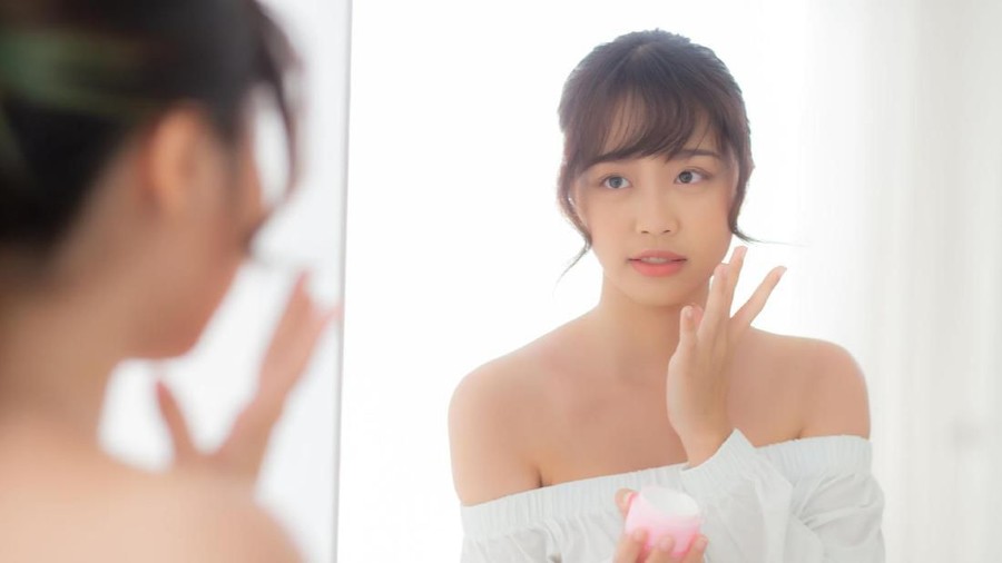 Beautiful portrait young asian woman applying cream moisturizer or lotion skin care cosmetic on face looking mirror, girl with treatment facial, health and wellness concept.
