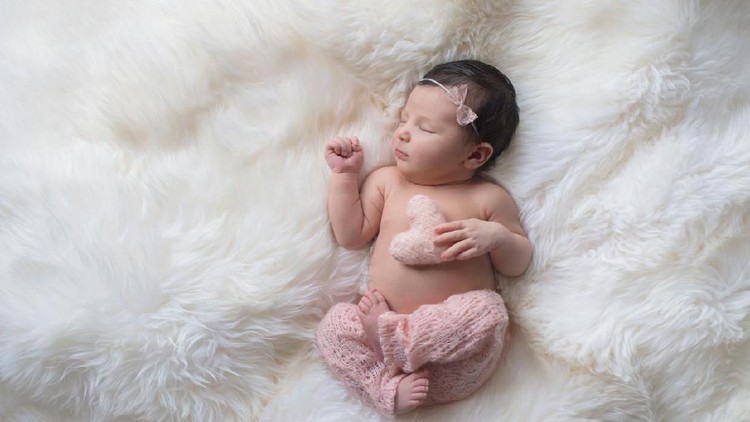 Sleeping, week old newborn baby girl wearing light pink, knitted pants and holding a tiny, heart shaped pillow. Shot in the studio on a white sheepskin rug.