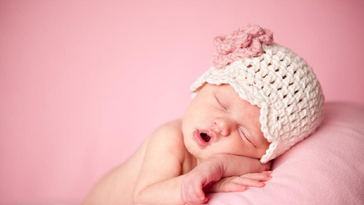 nama bayi Sleeping, week old newborn baby girl wearing light pink, knitted pants and holding a tiny, heart shaped pillow. Shot in the studio on a white sheepskin rug.