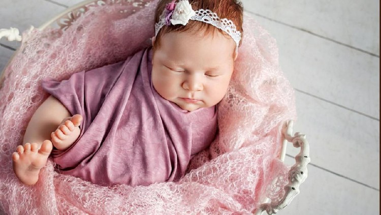 nama bayi Sleeping, week old newborn baby girl wearing light pink, knitted pants and holding a tiny, heart shaped pillow. Shot in the studio on a white sheepskin rug.
