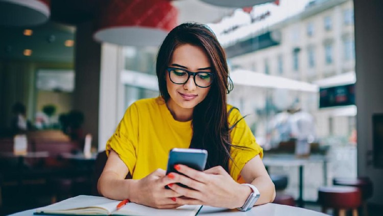 Young woman blogger in spectacles publishing new post on own website and installing new application for editing photos in social networks on digital smartphone device connected to free 4G internet