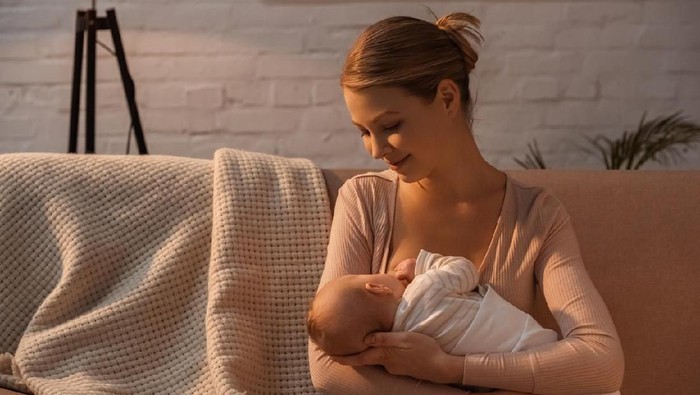 Confident female pediatrician examines a newborn baby. The baby's mother is holding her. The doctor is touching the baby's head. Baby bottles and a doctor's bag are on a table.