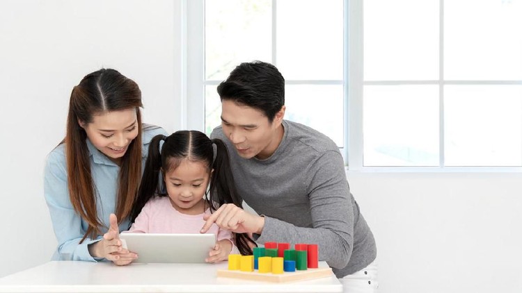 Happy young little asian cute girl watching or playing digital tablet, laptop or mobile with cheerful parents on desk in family time concept in white room with window. Preschool children learning.