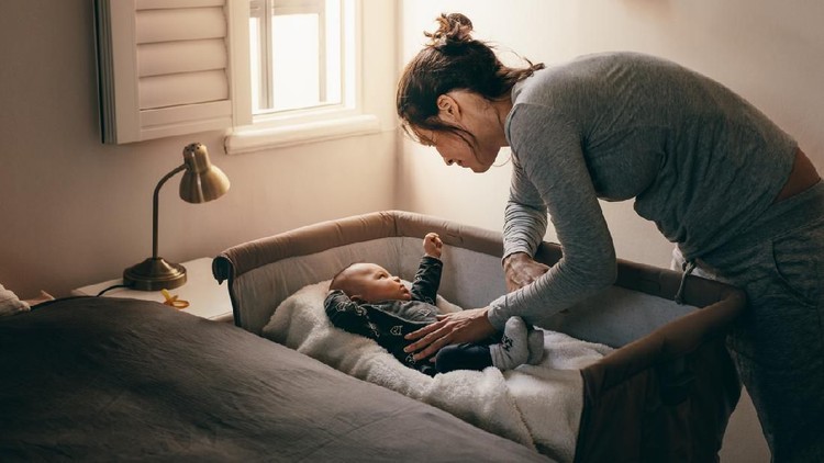 Mother putting her baby to sleep on a bedside baby crib. Woman bending forward over a crib to check her sleeping baby.