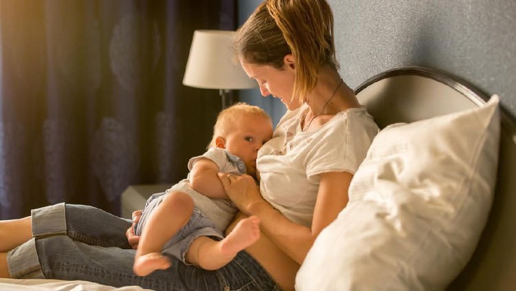 1126117897 Young mother, breastfeeding on sunset on quiet evening her little baby boy in sunny living room