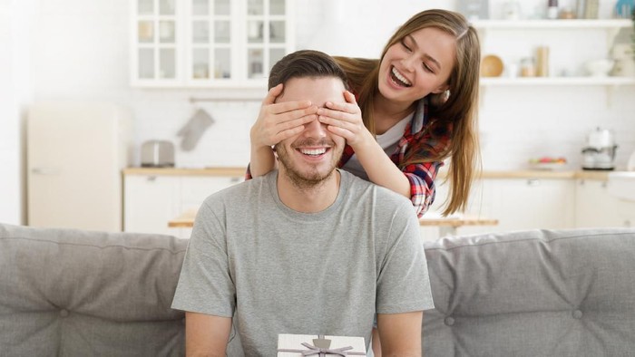 Young female keeps eyes closed to his boyfriend at home, while he is sitting on sofa with gift box, celebrating birthday