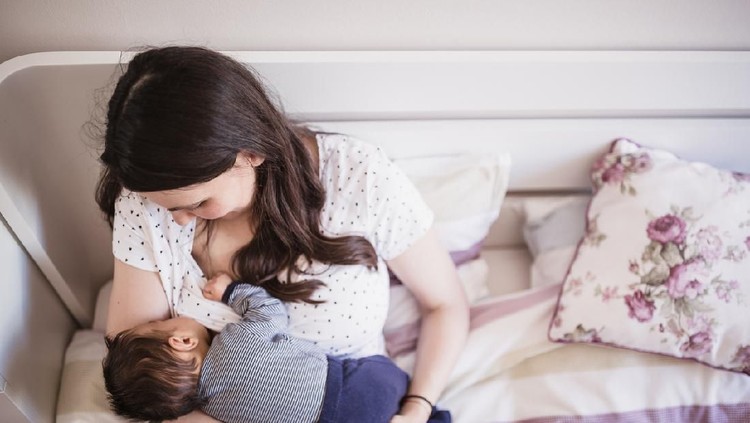 Mother and newborn baby boy are indoors in bedroom. Mother is breastfeeding her son while lying in bed.