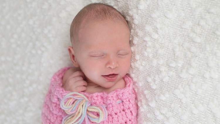 694153448 Headshot of a smiling three week old newborn baby girl bundled up in a light pink, crocheted, snuugle sack. She is lying on a white, bouncle blanket.
