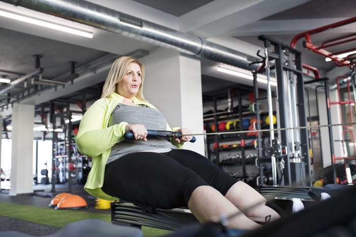 Attractive overweight woman in modern gym exercising on rowing machine. Chubby young blonde woman at fitness center.