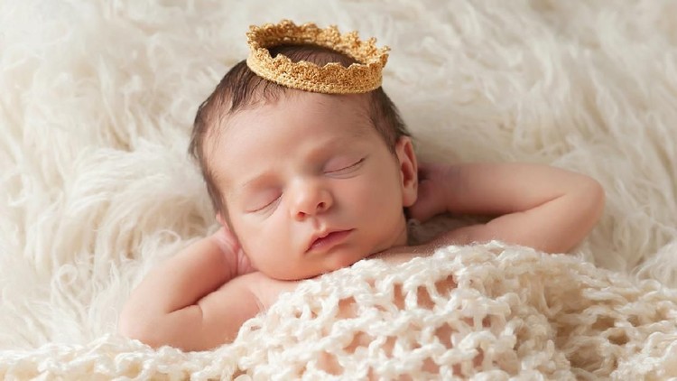 Portrait of a 12 day old newborn baby boy wearing a gold crown. He is sleeping on a beige flokati rug with his hands behind his head.
