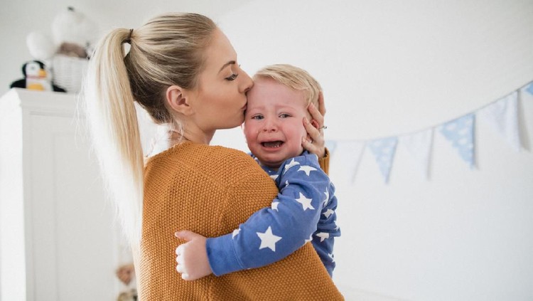 The little boy is crying in the crib and pulling his hands to his mom