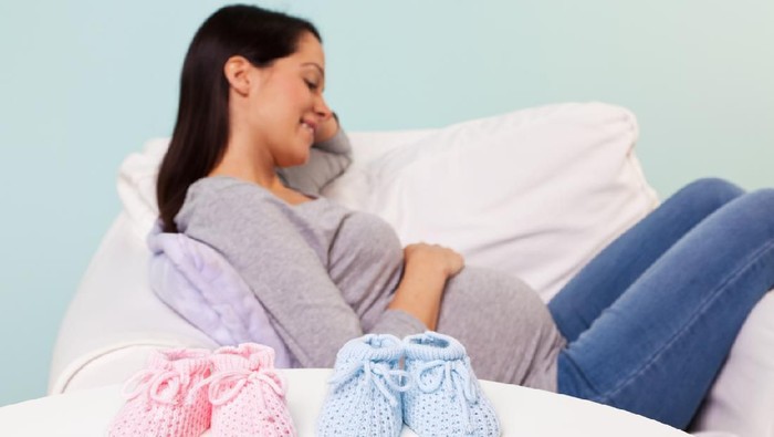 Photo of a pregnant woman at home sitting in an armchair withthe focus on a pair of boy and girl knitted baby booties on the table in front of her.