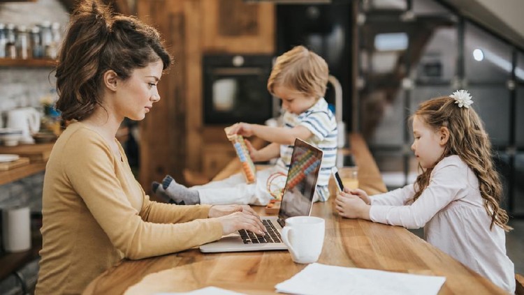 Young mother working on a computer at home while her small kids are around her playing.