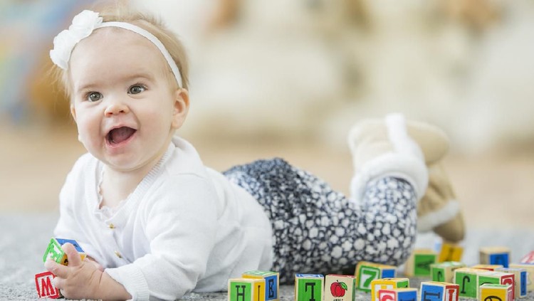 A Caucasian baby is indoors in a daycare. She is wearing a cute outfit. She is lying on the carpet and playing with colorful toy blocks.