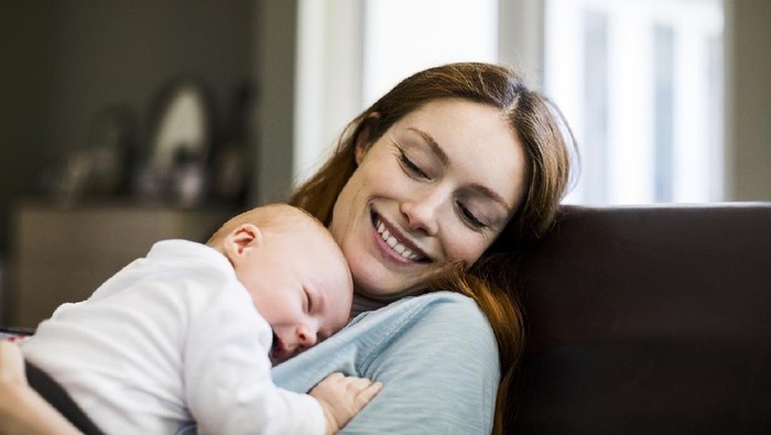Young mother, holding her sick toddler boy, breastfeeding him at home, sunny living room