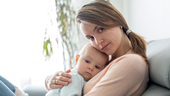 Young mother, holding her sick toddler boy, breastfeeding him at home, sunny living room