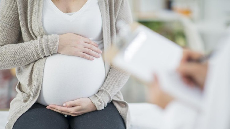 A pregnant Caucasian woman is indoors in a doctor's office. Her female doctor is wearing medical clothing. The woman is holding her stomach which the doctor writes on a clipboard.