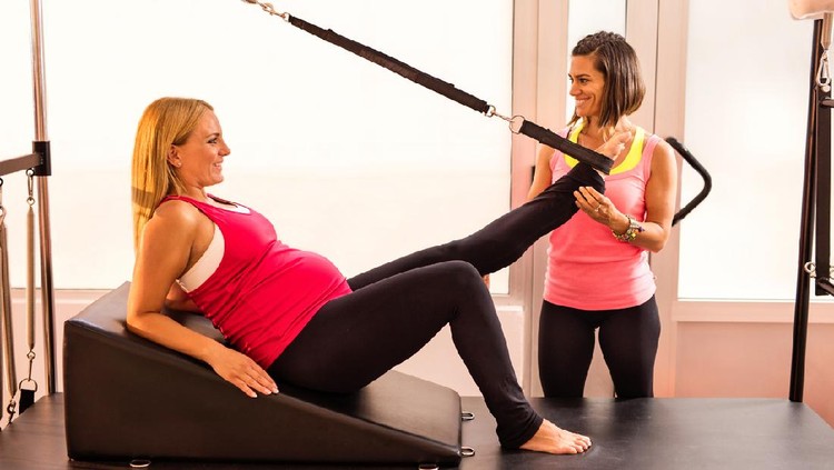 Smiling pregnant woman stretching her leg on reformer machine while Pilates trainer ins assisting her.