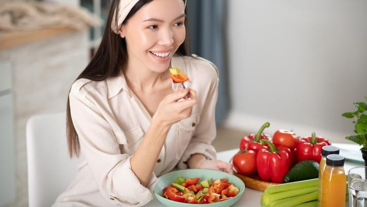 Right food. Good looking smiling young woman in a white blouse sitting at a table at home, eating with fork natural vegetable salad.
